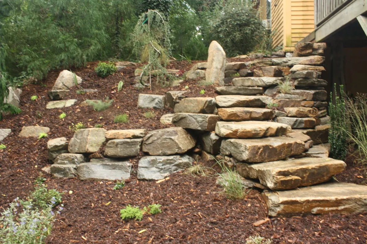 Terraced hillside landscaping with natural stone retaining walls, rustic stone steps, boulders, mulch beds, and woodland plantings